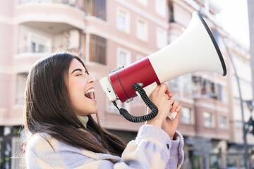 Young Brazilian woman at outdoors shouting through a megaphone to announce something in lateral position - obrazy, fototapety, plakaty