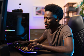 Professional male pro gamer sits comfortably in chair leaning over computer, playing video games, focused on coming up with strategy, room lit with purple blue led lights © ABCreative