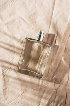 Transparent bottle of perfume on light grey fabric background. Fragrance presentation with daylight and palm leaves shadow. Trending concept in natural materials . Women's and men's essence.