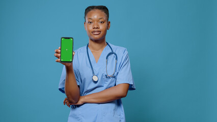 Portrait of medical assistant holding smartphone with green screen. Woman nurse showing device with isolated mockup template and chroma key background on display at camera in studio.