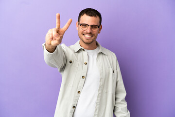 Brazilian man over isolated purple background smiling and showing victory sign