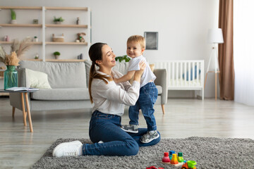 Pretty young mother enjoying time with her adorable toddler son at home, playing with kid boy in...