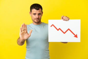 Brazilian man over isolated purple background holding a sign with a decreasing statistics arrow symbol and doing stop sign