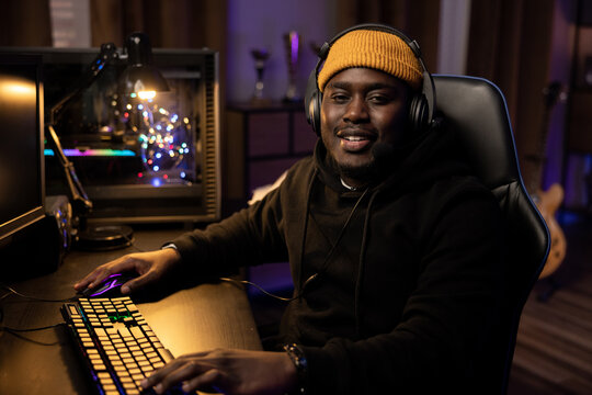 A dark room of a man sitting in front of a computer wearing a black sweatshirt and a yellow cap with a headset. Music producer's studio, rapper, singer processing song tracks. Guitar in the background