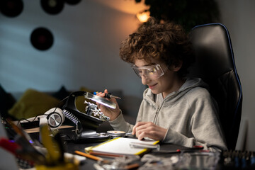Afro boy wearing safety glasses soldering motherboard in room at desk. The child is developing a hobby for electronics and is working on a school project.