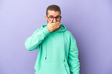 Young handsome caucasian man isolated on purple background covering mouth with hand