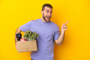 Young caucasian making a move while picking up a box full of things isolated on yellow background...