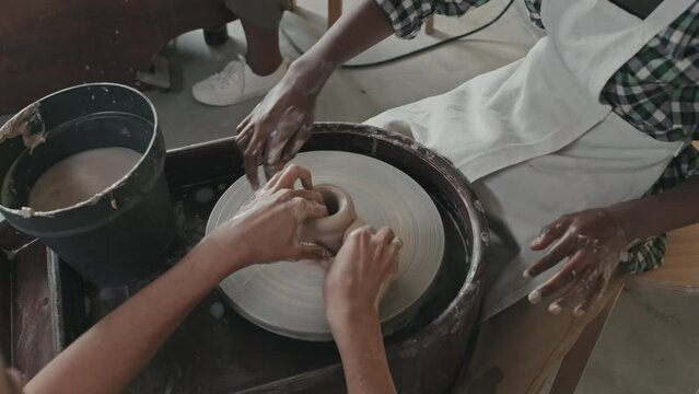High-angle of cropped Black boy sitting at pottery wheel and learning making crafts of clay in workshop at daytime, unrecognizable teacher helping student