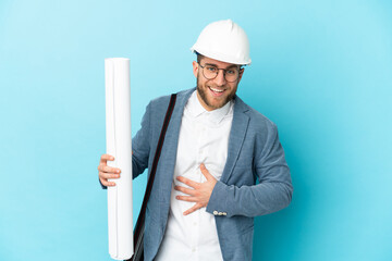 Young architect man with helmet and holding blueprints over isolated background smiling a lot