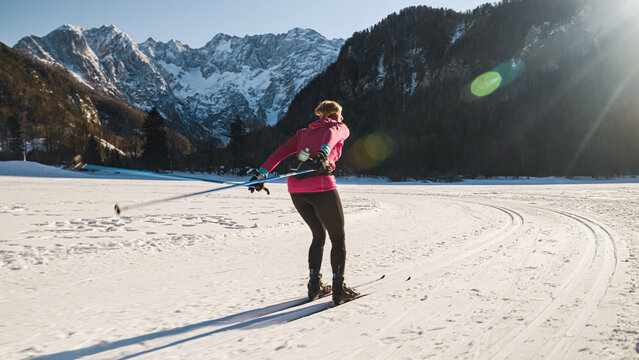 Caucasian woman cross country skiing on snowy flat terrain, at the foothill of beautiful mountain peaks, wide shot.
