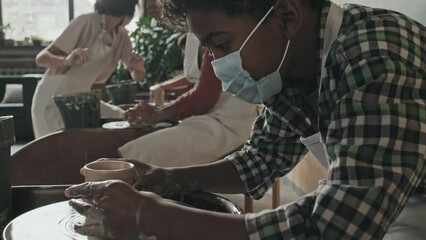 Low angle side view of little Black boy wearing apron and face mask, sitting and making craft of clay on pottery wheel in workshop at daytime - Powered by Adobe