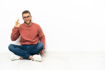 Young handsome man sitting on the floor showing ok sign with fingers