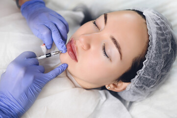 Obraz na płótnie Canvas Cosmetologist in beauty salon doing lip augmentation procedure with hyaluronic acid. The beautician pierces the lips with a needle. Young woman doing subcutaneous injection to increase lips shape.