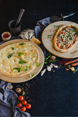 Photo of 2 pizzas on a textured black table in a restaurant. Top view. There are various ingredients nearby: cheese, mushrooms, tomatoes, spices, flour.