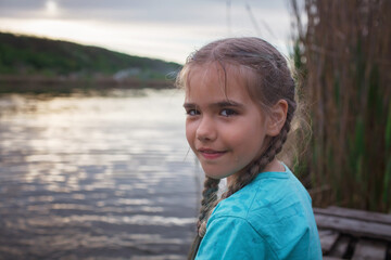 Portrait of happy little girl sitting on pontoon bridge on the bank of lake and enjoying warm sunny...