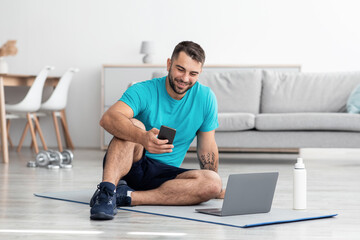 Happy smiling young caucasian guy sitting on mat on floor typing on phone and looking at computer...