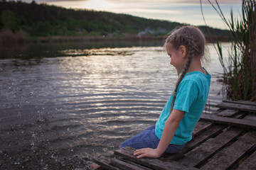 Happy little girl sitting on pontoon bridge on the bank of lake and enjoying warm sunny evening,...