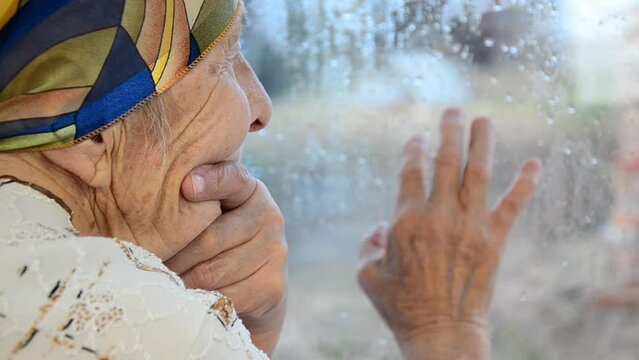 Grandmother looks out the window alone. Grandmother's wrinkled hands touch the window. Grandma draws on glass with her finger. Elderly woman is sad looking out the window.The concept of the elderly