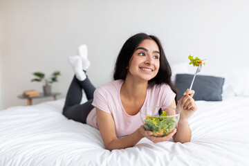 Lovely millennial Indian woman eating fresh vegetable salad on bed at home, blank space. Weight...