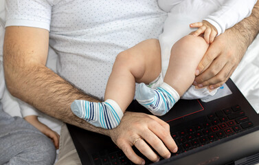 Naklejka premium cute little baby in father's arm is playing with his hands. father and son during pandemic, working online from home. black laptop on legs. boys are sitting on the bed for kids. fatherhood concept.