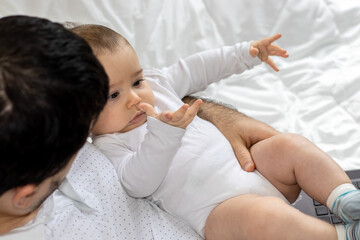 Fototapeta na wymiar cute little baby in father's arm is playing with his hands. father and son during pandemic, working online from home. black laptop on legs. boys are sitting on the bed for kids. fatherhood concept.