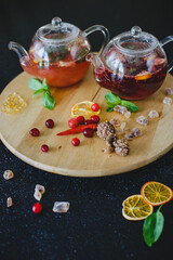Two teapots with fruit berry tea on a round textured wooden board on a black table in a restaurant. Next to sweets, candied fruits, berries