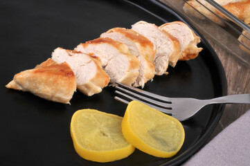 Blanc de poulet cuit coupé dans une assiette noire avec deux rondelles de citron et une fourchette
