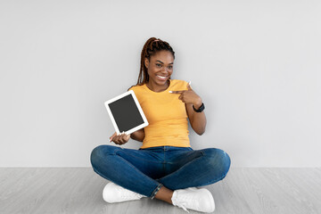 Black woman pointing at tablet with blank screen, sitting on floor against grey studio wall, mockup...