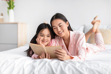 Happy glad chinese teenage girl and young lady in pink pajamas rest at home, looking at book