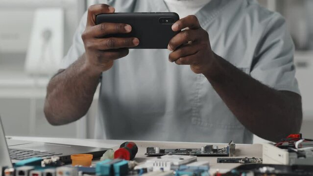 Tilt up shot of African American technician taking picture of electronic components with smartphone while working at desk in laboratory