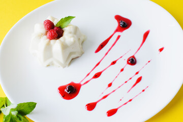 Mousse cake on a white plate with raspberries, mint and syrup on a yellow background