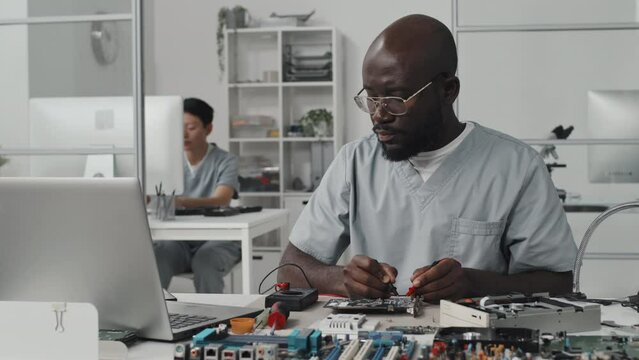 African American engineer testing electronic component with multimeter and working on laptop at desk in laboratory