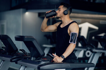 Fototapeta na wymiar Arab Guy Drinking Protein Cocktail From Sport Shaker While Training On Treadmill