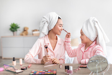 Cheerful chinese teenage girl and young female in pink pajamas and towels on heads do makeup in bedroom