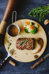 Photo of a grilled steak on a round wooden board in a restaurant. There are various appliances, vegetables, wine nearby