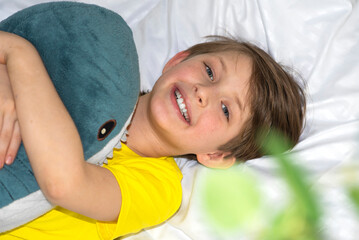 a cheerful european boy lies in a white bed and hugs a soft toy shark