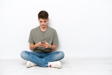 Teenager Russian man sitting on the floor isolated on white background sending a message with the mobile