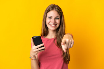 Young woman using mobile phone isolated on yellow background points finger at you with a confident...