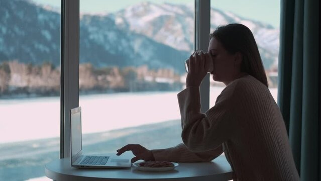 Woman freelancer working from home using laptop while sitting at a table by the window.