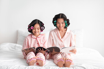 Smiling young asian woman and teen girl with curlers watching magazine and doing pedicure on bed