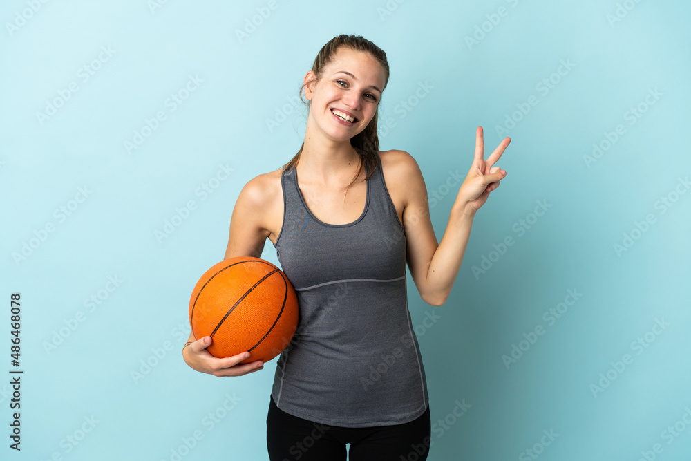 Canvas Prints Young woman playing basketball isolated on blue background smiling and showing victory sign