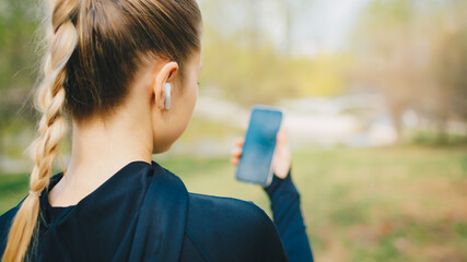 Young smiling girl making sport and running in the park using her phone to listen the music with wireless headphones on sunset in the city watching the screen