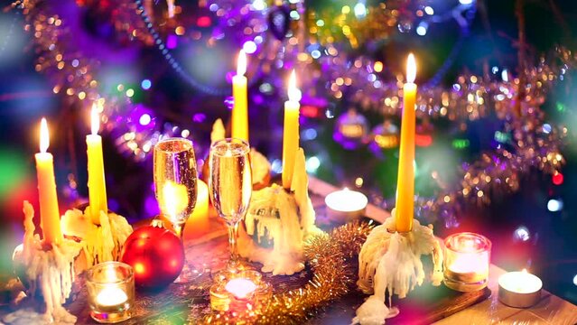 Close up of burning candles and two glass of champagne with funny bubbles against Christmas tree background twinking garland lights seamless looping footage selective focus.