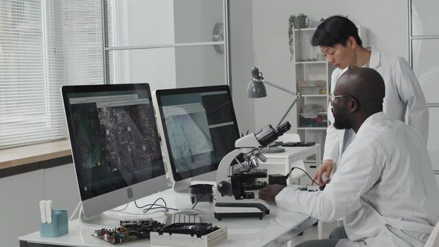 African American male engineer using microscope and discussing chip image on computer screen with Asian female colleague while working together in laboratory