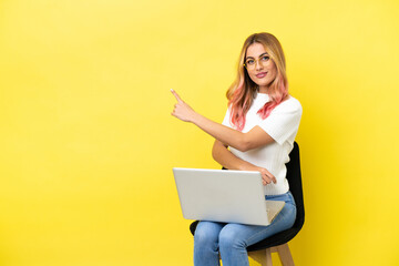 Young woman sitting on a chair with laptop over isolated yellow background pointing back - obrazy, fototapety, plakaty