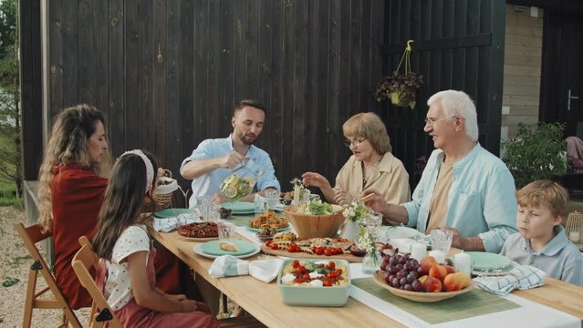 Tracking in of Caucasian family members of three generations sitting together at table in backyard of house on sunny summer weekend, having healthy lunch, smiling and chatting