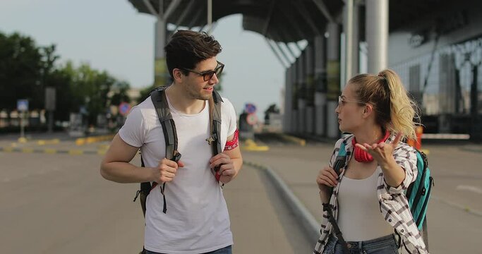 Couple Two Young Caucasian People Have Cheerful Conversation While Walking Near Airport. In Summer, Tourists Dressed Casual Clothes With Backpacks Over Their Shoulders. Tourism And Friendship Concept