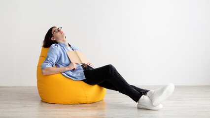 Remote education concept. Bored male student sleeping with book, sitting in beanbag chair, panorama, free space