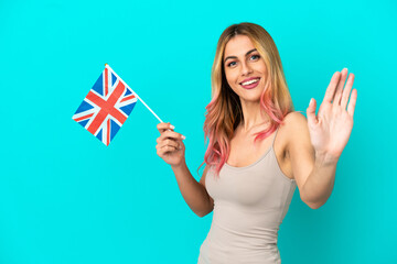 Young woman holding an United Kingdom flag over isolated blue background saluting with hand with...