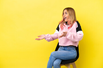 Young woman sitting on a chair over isolated yellow background with surprise expression while looking side